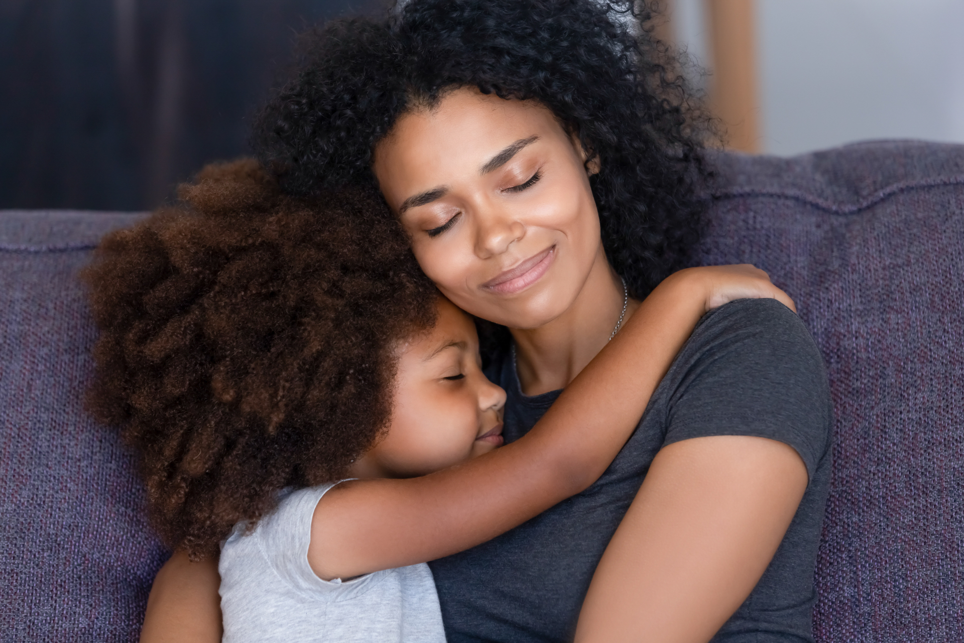 A mother holding her young daughter close on a couch, both with eyes closed and smiling